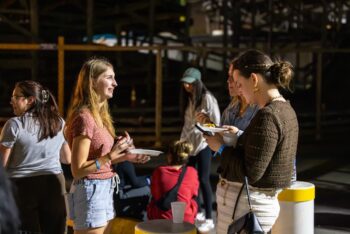 College students chat and share a meal during an evening gathering at Beach Reach, holding plates of food in a dimly lit open-air venue.