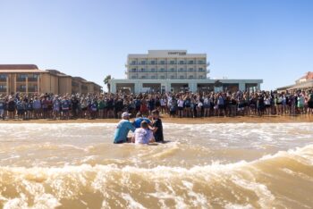 A person is baptized in the ocean surf as a large crowd of Beach Reach participants lines the shore, cheering and celebrating with a beachfront hotel visible in the background.