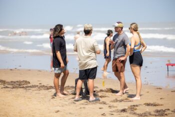 A small group of young adults stand barefoot on the beach near the shoreline, talking together on a sunny day during Beach Reach.