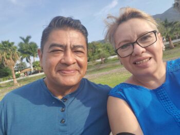 River Ministry missionary Pedro Muñoz and his wife smile in a selfie outdoors in Reynosa, Mexico, with palm trees and mountains in the background